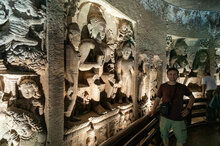 Eine Innenansicht einer Höhle mit kunstvoll gemeißelten Reliefs, die Buddha und andere Figuren darstellen, beleuchtet von weichem Licht. Eine Person steht im Vordergrund und betrachtet die beeindruckenden Schnitzereien.