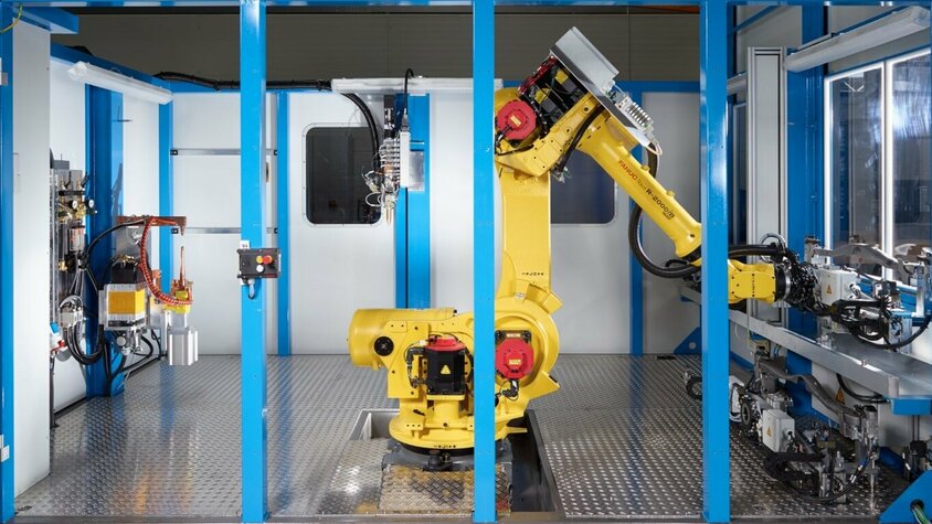 Eine moderne Fügetechnik-Anlage mit einem gelben FANUC-Roboterarm in einem abgeschirmten Arbeitsbereich. Der Roboter ist zentral in der Anlage positioniert und bereit für automatisierte Klebeaufgaben. Umgeben von blauen Rahmenstrukturen zeigt die Szene eine hochmoderne Fertigungsumgebung mit präzisen mechanischen Komponenten und fortschrittlichen Steuergeräten. Diese Anlage steht für Präzision, Effizienz und Automation in der Fügetechnik.