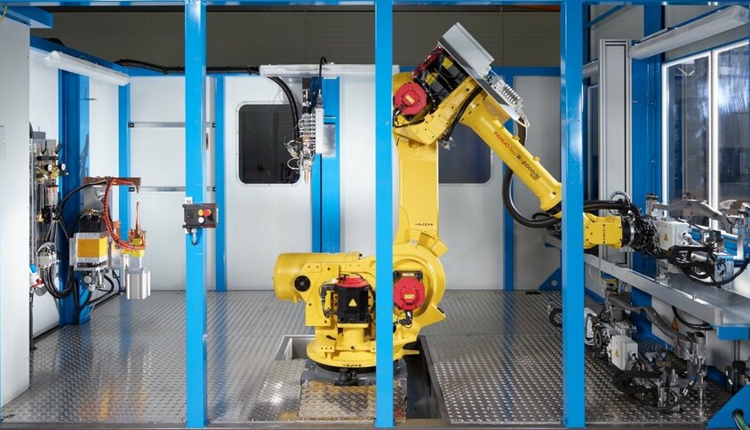 Moderní systém spojovací techniky se žlutým robotickým ramenem FANUC ve stíněném pracovním prostoru. Robot je umístěn uprostřed systému a je připraven k automatickému spojování. Scéna obklopená modrými rámovými konstrukcemi ukazuje nejmodernější výrobní prostředí s přesnými mechanickými součástmi a pokročilými řídicími jednotkami. Tento systém je ztělesněním přesnosti, efektivity a automatizace v technologii spojování.