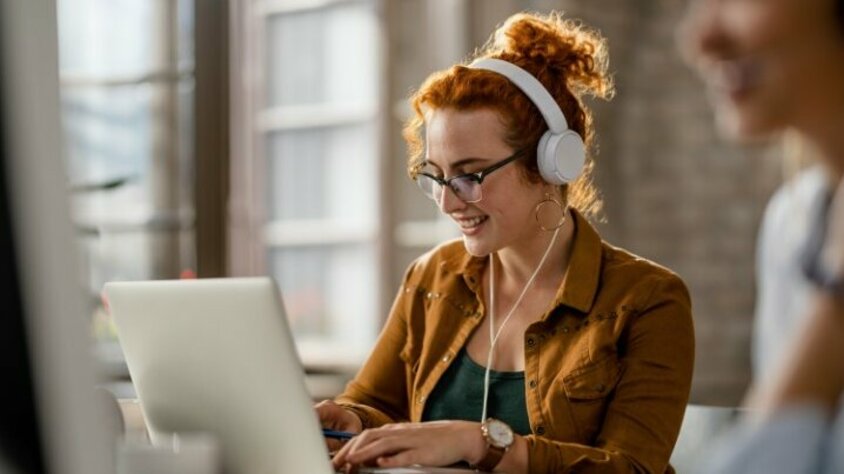 Lächelnde kreative junge Frau hört Musik mit Kopfhörern, während sie im Büro an einem Computer bei ihrem Praktikum Job in Steyr bei weba arbeitet.