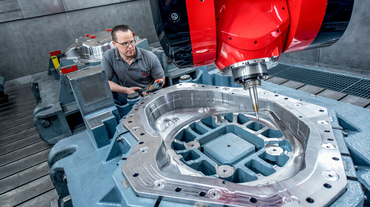 Ein Facharbeiter bedient eine hochmoderne CNC-Fräsmaschine mit einem großen Werkzeugbauteil. Die glänzende Metalloberfläche des Bauteils zeigt präzise Zerspanungsergebnisse, während die rote Fräseinheit prominent im Vordergrund zu sehen ist.