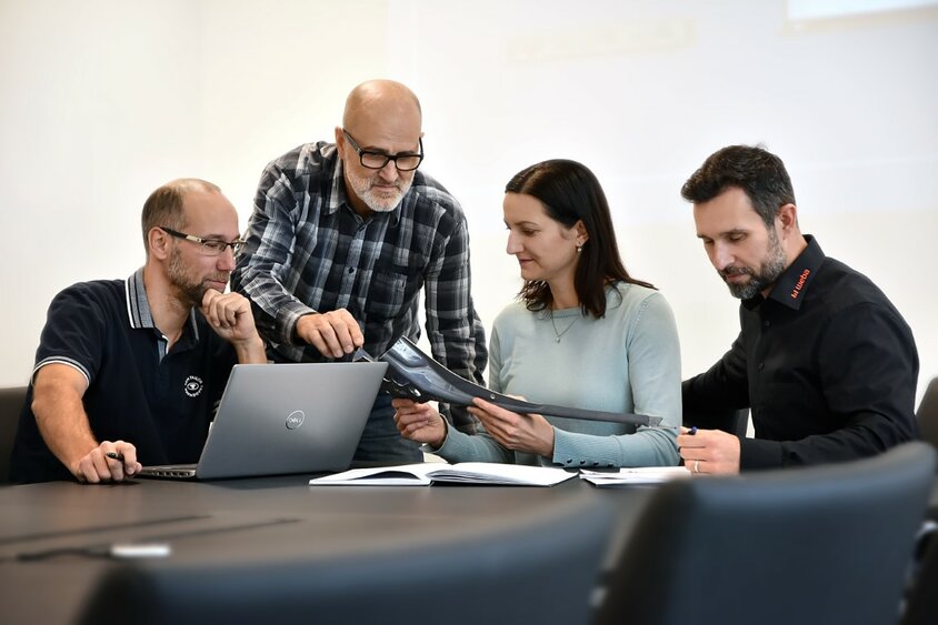 Drei männliche und eine weibliche Mitarbeiterin von weba Werkzeugbau sitzen in einer Besprechung über ein Stahlbauteil. Ein Laptop und Unterlagen liegen auf dem Tisch.