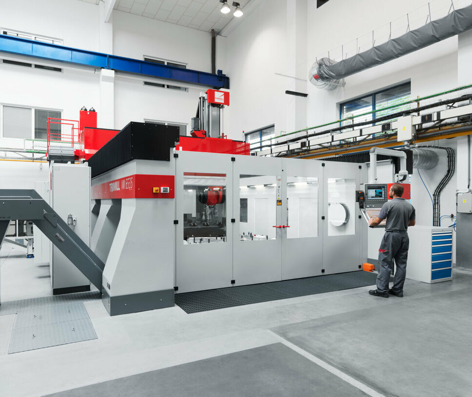 A machine operator is working at a modern CNC machining center for mechanical machining. The machine is installed in a bright and clean production environment and features an advanced control panel that allows for precise operation. The robust construction of the machine and its enclosed cabin indicate high-precision machining processes. To the left of the machine, there is a material feeder that supports the smooth workflow of the machining process.