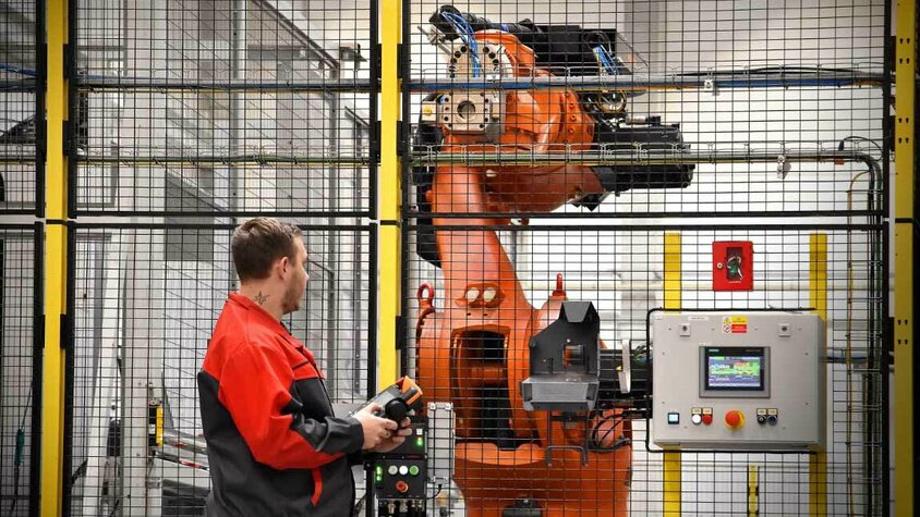 Ein Mitarbeiter in roter Arbeitskleidung bedient einen Industrieroboter in einer gesicherten Produktionszelle. Der Roboterarm ist hinter einem Sicherheitszaun sichtbar, und der Bediener hält ein Steuergerät in der Hand, um die Bewegungen des Roboters zu überwachen oder anzupassen. Neben dem Roboter befindet sich ein Bedienfeld mit einem digitalen Display, das zur Kontrolle und Überwachung der Roboterprozesse dient. Die Szene zeigt den Einsatz von Automatisierung und fortschrittlicher Robotik in der mechanischen Bearbeitung.