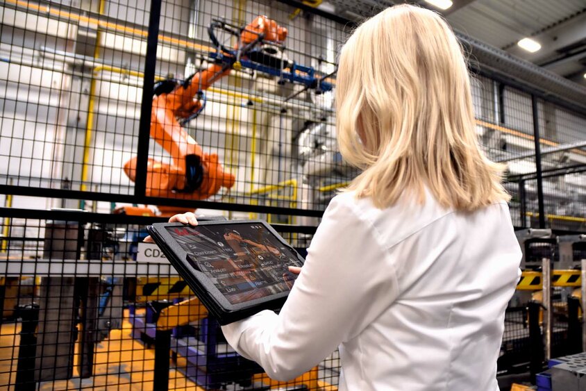 Eine Frau steht in einer industriellen Umgebung bei weba Werkzeugbau vor einem großen Robotergreifer einer Warmumformlinie. In ihren Händen hält sie ein Tablet, auf dem sie Daten überwacht oder Steuerbefehle für den Roboter eingibt. Der Fokus liegt auf der Interaktion zwischen moderner Technologie und menschlichem Eingreifen.