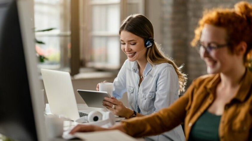 Lächelnde weibliche Person mit Headset, die Kaffee trinkt und im Büro das Touchpad nutzt. Das Bild symbolisiert glücklichen Berufseinstieg in Steyr bei weba.