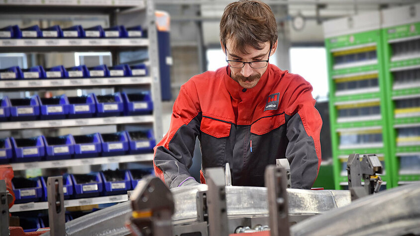 Ein Werkzeugbautechniker baut konzentriert ein Presswerkzeug für anschließenden Werkzeug-Tryout und die Umformung von Strukturbauteilen zusammen; Im Hintergrund sind unscharf Regale mit Hilfs- und Betriebsstoffen 