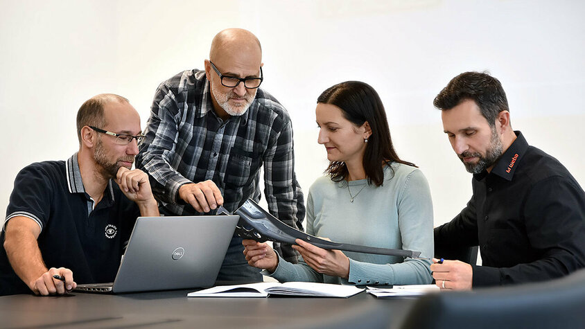 Vier Personen sitzen an einem Besprechungstisch: Eine Person arbeitet an einem Laptop, eine andere betrachtet aufmerksam Unterlagen, während zwei weitere Personen sich angeregt über ein Stanzteil unterhalten. Die Szene zeigt eine konzentrierte und engagierte Arbeitsatmosphäre. Berufserfahrene und Profis finden bei weba ihren Traumjob in Steyr. 