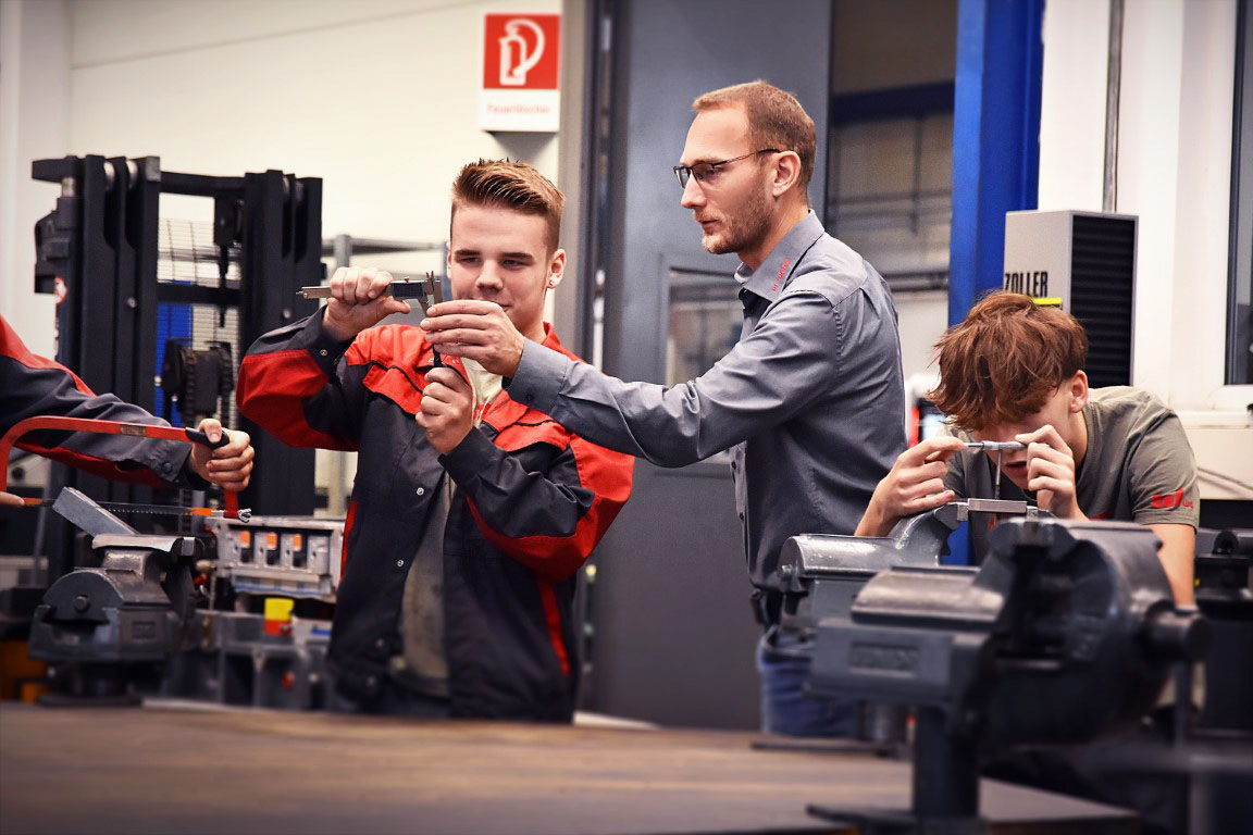 Ein Lehrling in der Werkzeugbautechnik, konzentriert beim Messen eines Stahlklotzes mit einem Messschieber, während der Ausbilder neben ihm steht und etwas erklärt und zeigt. Daneben spannt ein weiterer Lehrling aufmerksam einen Stahlklotz in einen Spannbock, was die praktische und lehrreiche Umgebung der Ausbildung verdeutlicht. Das Bild ist Symbolfoto für die Lehre in Steyr in der Werkzeugbautechnik Modul H6 aus der Metalltechnik bei weba Werkzeugbau.